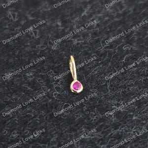 Puede incluir: Un pequeño colgante dorado con una gema rosa brillante en forma de lágrima. El colgante tiene un lazo para una cadena. El fondo es una superficie oscura y texturizada.