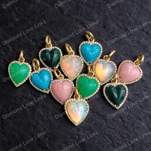 May include: A collection of heart-shaped pendants in various colors, including green, turquoise, pink, and opal. Each pendant has a gold-colored frame and is encrusted with small, clear stones. The pendants are displayed on a dark surface.