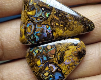 Puede incluir: Dos piedras preciosas de ópalo pulidas. Los ópalos muestran un mosaico de colores, incluyendo amarillo, marrón, azul y destellos iridiscentes. El ópalo superior es triangular y el inferior tiene forma de corazón. Los ópalos tienen un aspecto texturizado y orgánico.