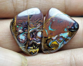 Puede incluir: Dos piedras preciosas de ópalo pulidas. Los ópalos tienen un color base marrón con destellos iridiscentes de azul, verde y amarillo. Los bordes están delineados con una matriz dorada, y las piedras tienen una forma suave y redondeada.