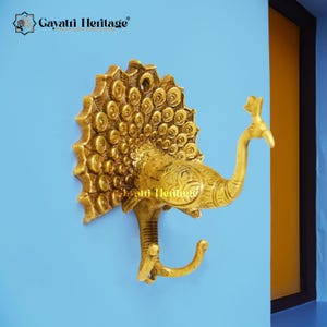 May include: A brass peacock wall hook with intricate detailing. The hook is shaped like a peacock with its tail fanned out. The hook is mounted on a light blue wall. The text "Gayatri Heritage" is visible at the top.