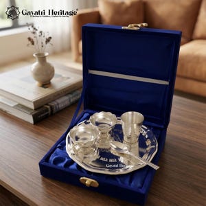May include: A silver puja set in a deep blue velvet box. The set includes a tray, three small bowls, a cup, and a spoon. The tray and bowls have a decorative floral design. The box is open, revealing the contents.