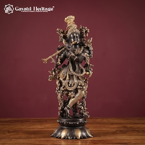 Puede incluir: Una estatua de metal en tono bronce oscuro de Krishna tocando una flauta, sobre una base en forma de loto. La figura presenta tallas intrincadas, joyas ornamentadas y una corona alta. El texto "Gayatri Heritage" es visible en la esquina superior.