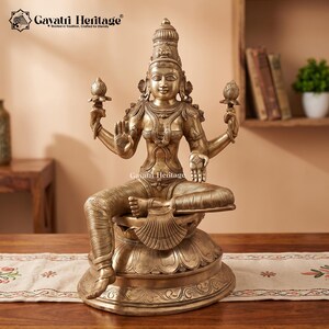 Può includere: Una statua in ottone di una divinità seduta su una base di loto, con quattro braccia, due mani che reggono boccioli di loto, una mano in gesto di benedizione e una mano in gesto di offerta, decorata con gioielli elaborati e una corona alta.