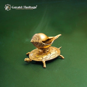 May include: A golden brass decorative item featuring a conch shell-shaped vessel atop a turtle-shaped base. The item has intricate detailing and is set against a green background. The text "Gayatri Heritage" is visible in the upper left corner.