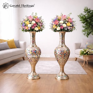 Puede incluir: Dos jarrones altos y ornamentados de latón con arreglos florales coloridos. Los jarrones tienen intrincados diseños florales y están colocados en una habitación bien iluminada. El texto "Gavatiri Heritage" es visible en la parte superior.