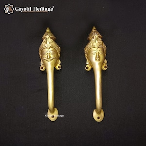 Peut inclure: Deux poignées de porte en laiton doré, chacune avec un motif de visage détaillé. Les poignées ont une forme longue et incurvée avec un visage décoratif en haut. Ces poignées sont probablement pour une armoire ou une porte, et sont en métal.
