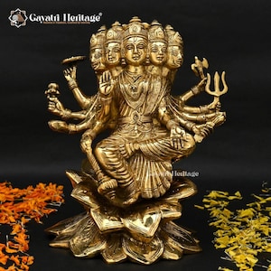 May include: A golden brass statue of a multi-faced deity seated on a lotus flower. The statue has multiple arms, each holding a different object. The deity is adorned with intricate details and sits against a dark background. The statue is a representation of Indian culture.