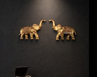 May include: Two golden elephant wall sculptures with raised trunks and ornate detailing. The elephants face each other, mounted on a dark wall. The brand name "Gayatri Heritage" is visible in the upper left corner.