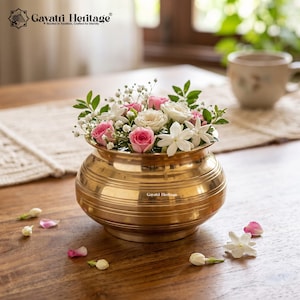 May include: A polished brass pot with horizontal ridged detailing, filled with a floral arrangement of pink roses, white jasmine flowers, and small white baby's breath. The text Gayatri Heritage is printed on the side of the brass pot.