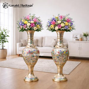Pode incluir: Dois vasos altos e ornamentados de latão com arranjos florais. Os vasos apresentam um padrão floral detalhado e colorido e são preenchidos com flores rosa, amarelas e azuis. Estão numa sala luminosa com um sofá e um armário. O texto "Gavatiri Heritage" é visível.