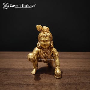 May include: A gold-toned metal figurine of a crawling child deity, featuring intricate jewelry, a peacock feather in the hair, and a small spherical object held in the right hand. The figure is depicted in a kneeling posture with detailed facial features.