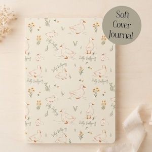 May include: A soft cover journal with a cream background featuring a repeating pattern of ducks, small yellow flowers, and hearts. The text "Dilly Dallying" is printed in a cursive script throughout the design. The cover has a matte finish.