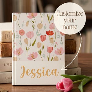 Puede incluir: Un diario personalizado con un diseño floral. La portada es crema con flores rosas, rojas y melocotón. El nombre "Jessica" está impreso en oro. También se ve el texto "Customize your name".