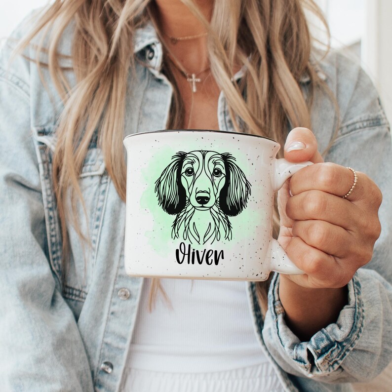 Puede incluir: Taza de cer&aacute;mica blanca con borde negro y dise&ntilde;o moteado. La taza presenta un dibujo lineal en blanco y negro de un perro con orejas largas, sobre un fondo aguamarina. El nombre "Oliver" est&aacute; escrito debajo.