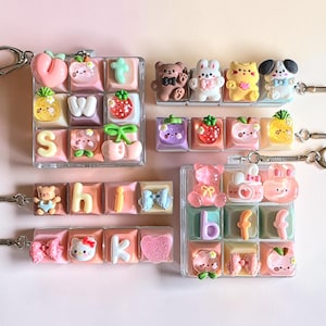 May include: A collection of keycap keychains featuring various pastel-colored square keys decorated with miniature resin figures including bears, rabbits, cats, fruits, bows, and letters. Some keychains are square grids while others are linear strips.