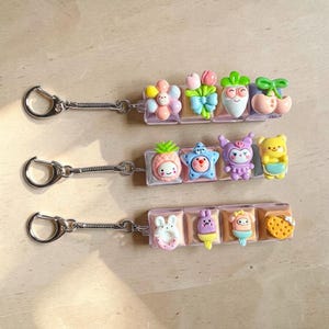 May include: Three keychains with decorative charms. Each keychain has a silver clasp and spring coil. Charms include a flower, tulips, a smiling carrot, cherries, a pineapple, a star, a purple character, a bear, a donut, ice cream, a pig, and a strawberry.
