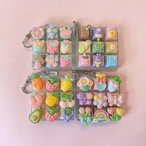 May include: Four square keychains featuring a grid of colorful, raised resin charms. The charms include miniature fruits, flowers, sweets, animals, and a rainbow with the word HAPPY. Each keychain has a silver metal ring attached to the corner.