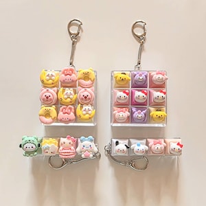 以下が含まれることがあります： 3x3のキャラクターキーキャップグリッドを備えた2つの正方形キーホルダーと、4つのキーキャップが並んだ2つの長方形キーホルダー。パステルカラーの様々な漫画のキャラクター、動物、食べ物のデザインが透明なベースの上に配置されています。