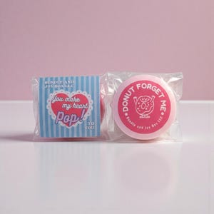 May include: Two wrapped pink treats with text. One has a blue and white striped background with a heart that says "You make my heart Pop!" The other says "Donut Forget Me" with a donut graphic.