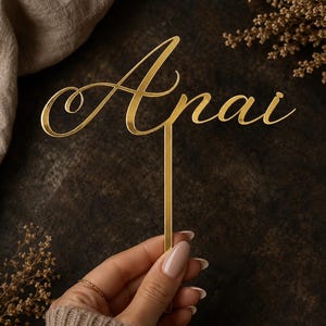 May include: A gold-colored cake topper featuring the name Anai written in a cursive script font. The topper has a reflective, smooth surface and includes a single vertical stem extending downward from the center of the lettering for insertion.