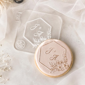Peut inclure: Un tampon à biscuits en acrylique transparent présentant un cadre hexagonal avec les lettres J & A et des détails floraux, à côté d'un biscuit rond rose clair gaufré avec le même motif hexagonal, des détails floraux et les lettres J & A.