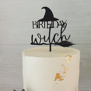 May include: A white frosted cake with gold leaf detailing, topped with a black cake topper reading "Birthday Witch" with a witch hat and broom design. A black cat decoration is at the base of the cake.