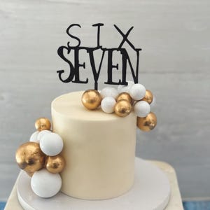 Puede incluir: Un pastel crema de dos niveles con decoraciones esféricas doradas y blancas. Un adorno negro para pastel dice "SIX SEVEN". El pastel está sobre un soporte de mármol y madera, perfecto para una celebración de cumpleaños.