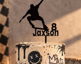 May include: A cylindrical cake decorated with black paint splatters, a skateboard, a smiley face, and the name Jaxson. A black cake topper features a silhouette of a person skateboarding with the number 8 and the name Jaxson.