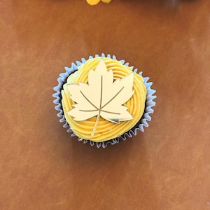 Peut inclure: Un cupcake avec un glaçage jaune et une décoration en forme de feuille d'érable dorée. Le cupcake est dans une caissette en papier blanche et noire. Le fond est une surface marron avec des tournesols et des ornements dorés.