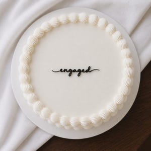 May include: A round white cake with smooth icing and a decorative piped border of white frosting around the edge. The centre of the cake features a black script topper with the word engaged written in a cursive font, positioned on the flat surface.