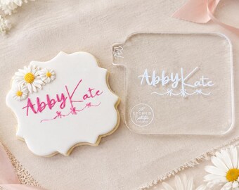 May include: A white sugar biscuit with scalloped edges, decorated with three small white daisy icing flowers and the pink script text "Abby Kate" above three small pink bows. Beside it is a clear acrylic stamp featuring the same text and design.