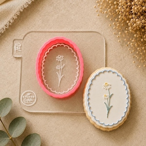 May include: A pink daisy-shaped cookie cutter and a decorated cookie. The cookie cutter is oval with a scalloped edge and a daisy design. The cookie is oval with a white icing daisy design and a blue border. The text "ETCHED Confection" is visible.