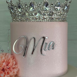 May include: A pink cake with the name "Mia" in silver script. The cake is topped with a silver crown adorned with large, clear gemstones. A peach-colored flower is on the side of the cake.