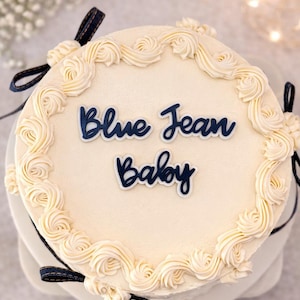 May include: A round cake with white frosting and decorative swirls around the edge. The cake has the words "Blue Jean Baby" in dark blue script. Dark blue denim ribbons are tied around the base of the cake.