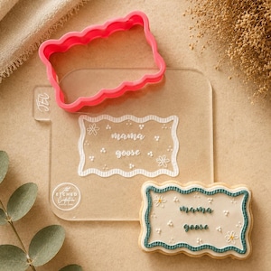 May include: A pink, scalloped-edge cookie cutter and a clear acrylic stamp with the words "mama goose" and floral accents. A decorated cookie with the same design is also present, featuring a teal border and white icing.