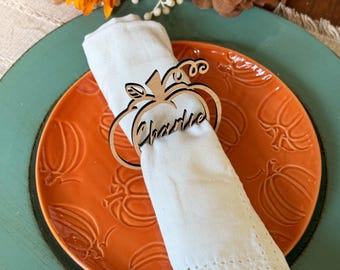 May include: A wooden pumpkin napkin ring with the name "Charlie" engraved, holding a white napkin. The napkin sits on an orange plate with pumpkin patterns, placed on a teal charger. The word "thankful" is visible in the background.