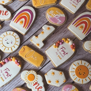 May include: Assortment of decorated sugar cookies with a birthday theme. Designs include smiling suns, rainbows, the number one, and the phrases "One", "First trip around the sun", and "Happy Birthday". The cookies are decorated with orange, pink, and white icing.