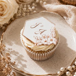 May include: A cupcake topped with white frosting and a hexagonal acrylic topper. The topper features the letters J & A in a copper-colored script font, accompanied by a delicate copper-colored floral line art illustration on a white background.