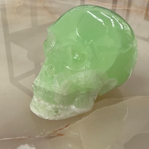 Puede incluir: Una escultura de cráneo tallada en piedra verde claro translúcida con inclusiones minerales blancas visibles. El objeto presenta una superficie lisa y pulida con contornos anatómicos detallados de las cuencas de los ojos, la cavidad nasal y la mandíbula.