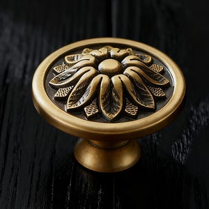 Può includere: Pomello per armadio in ottone in stile antico con design floreale. Il pomello presenta una faccia circolare con un motivo floreale in rilievo e una base corta e arrotondata. La finitura è di un caldo colore ottone anticato, su una superficie di legno scuro.
