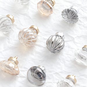 Puede incluir: Un surtido de tapones decorativos de vidrio para botellas con tapas doradas. Los tapones varían en forma y color, incluyendo tonos claros, esmerilados y ámbar. Los tapones están dispuestos sobre una superficie blanca texturizada.