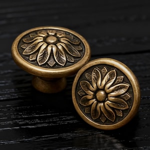 May include: Two antique brass cabinet knobs with a floral design. The knobs are round with a raised flower pattern and a dark, aged finish. The knobs are set against a dark wood surface.