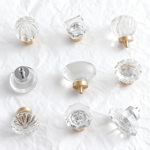 Könnte beinhalten: Neun transparente Glasschrankknöpfe in verschiedenen Formen, darunter runde, ovale und facettierte Designs. Jeder Knopf hat einen goldfarbenen Sockel und eine silberfarbene Schraube. Die Knöpfe sind auf einer weißen, strukturierten Oberfläche angeordnet.