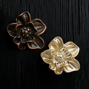 May include: Two decorative flower-shaped cabinet knobs. One is bronze-colored, the other is gold. The knobs have detailed petal and center designs. They are set against a dark, textured background, suggesting a rustic or vintage aesthetic.