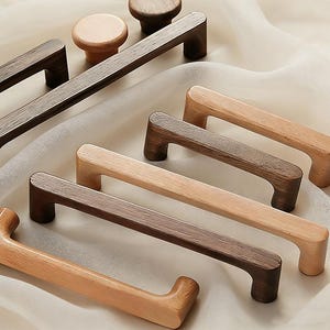 May include: A collection of wooden cabinet hardware, including knobs and pull handles. The knobs are round, and the handles are rectangular with curved grips. The wood tones vary from light to dark brown, showcasing natural grain patterns.