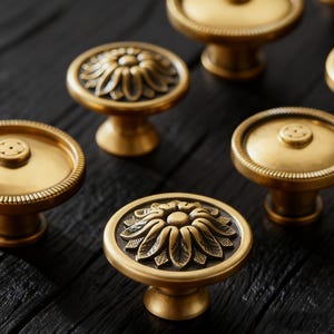 May include: Close-up of several antique brass cabinet knobs. The knobs have a variety of designs, including a floral pattern and a circular design with a beaded edge. The knobs are on a dark wooden surface.