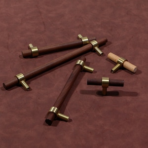 May include: A selection of wooden cabinet pulls with brass detailing. The pulls feature a cylindrical design with a dark brown finish, paired with gold-toned brass hardware. One pull is made of light-coloured wood. The pulls are displayed on a brown surface.