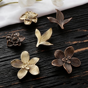 May include: A collection of decorative cabinet knobs in various floral and leaf designs. The knobs are made of metal and feature detailed textures and finishes in gold and bronze tones. The knobs are arranged on a dark wood surface.