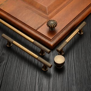 May include: A collection of antique brass cabinet hardware, including two long bar pulls, two smaller pulls, and two round knobs. The hardware features a ribbed design and is set against a wooden cabinet.